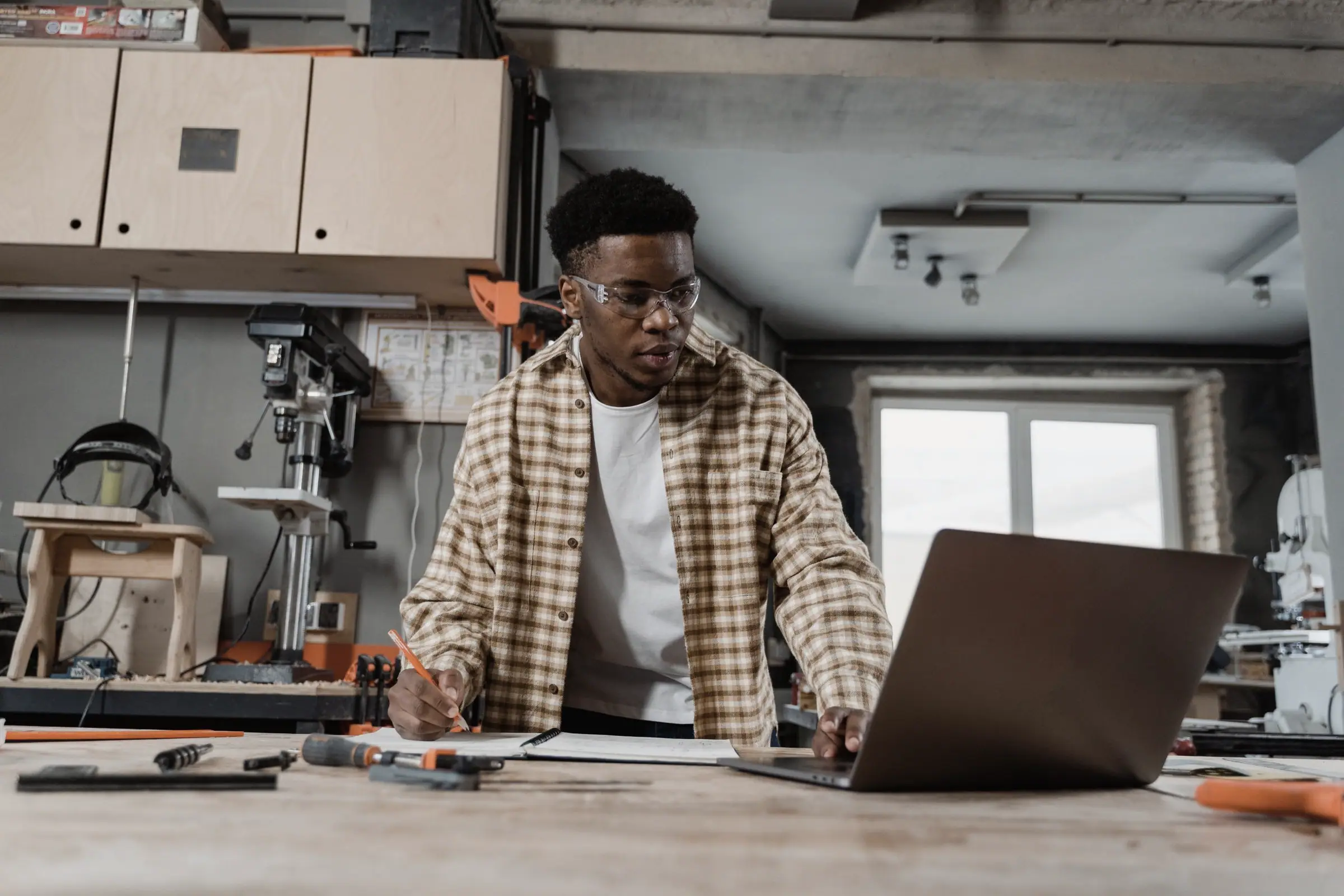 A man in safety glasses at a workshop bench, working on a laptop.